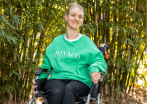 Janna Harrison, wearing a green jumper, sitting in her wheelchair, with green trees behind her.